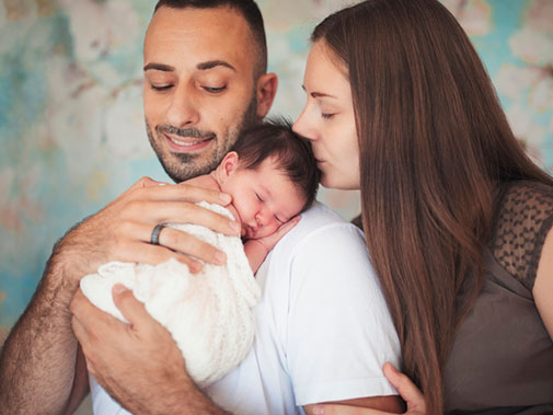 Father holding sleeping newborn on his shoulder while the mother kisses the baby on the forehead